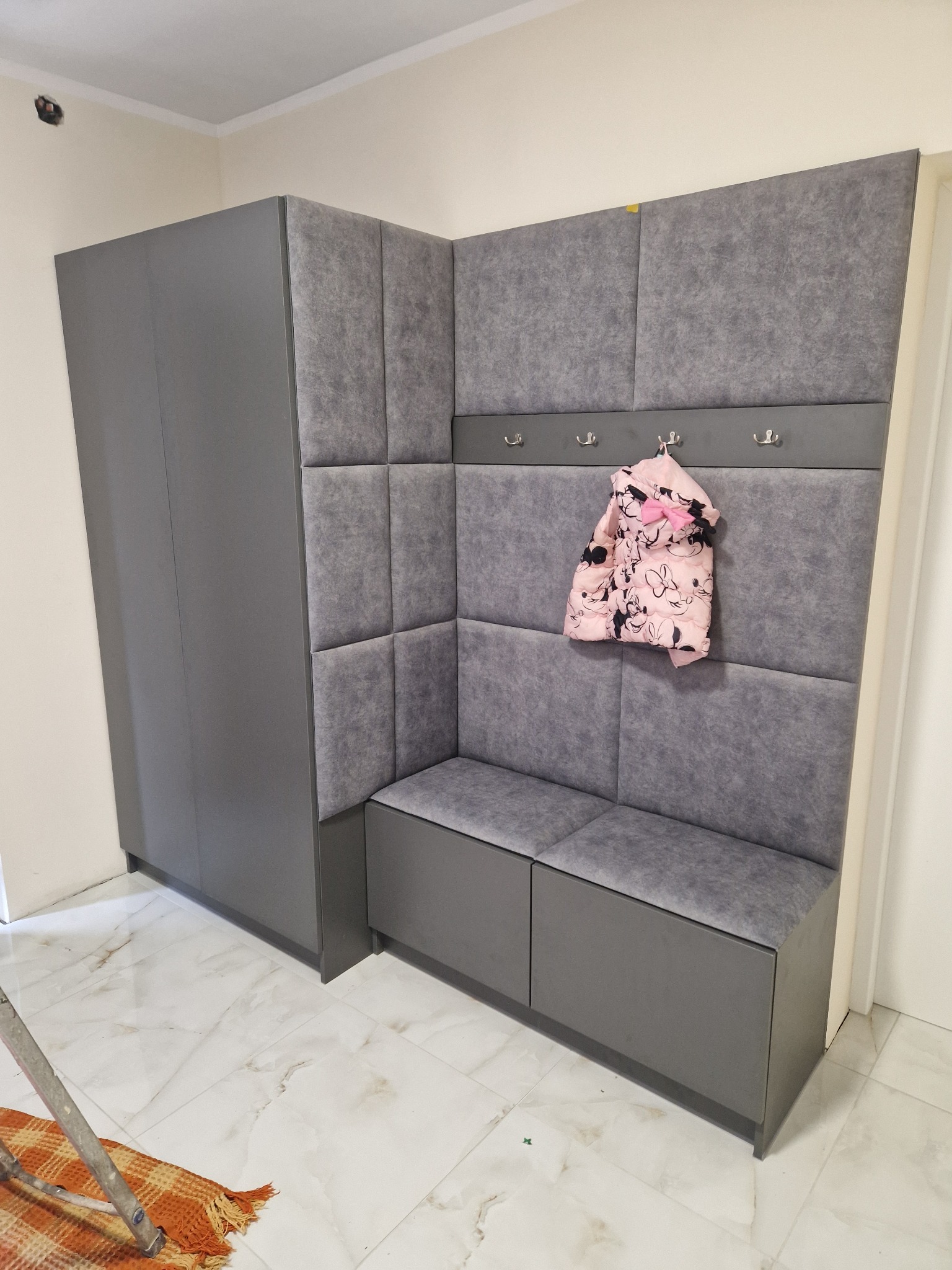 Szara zabudowa przedpokoju z szafą, siedziskiem i panelem tapicerowanym, z zawieszoną kurtką dziecięcą w kwiaty na wieszaku, widoczne fragmenty drabiny i podłogi z płytek imitujących marmur.