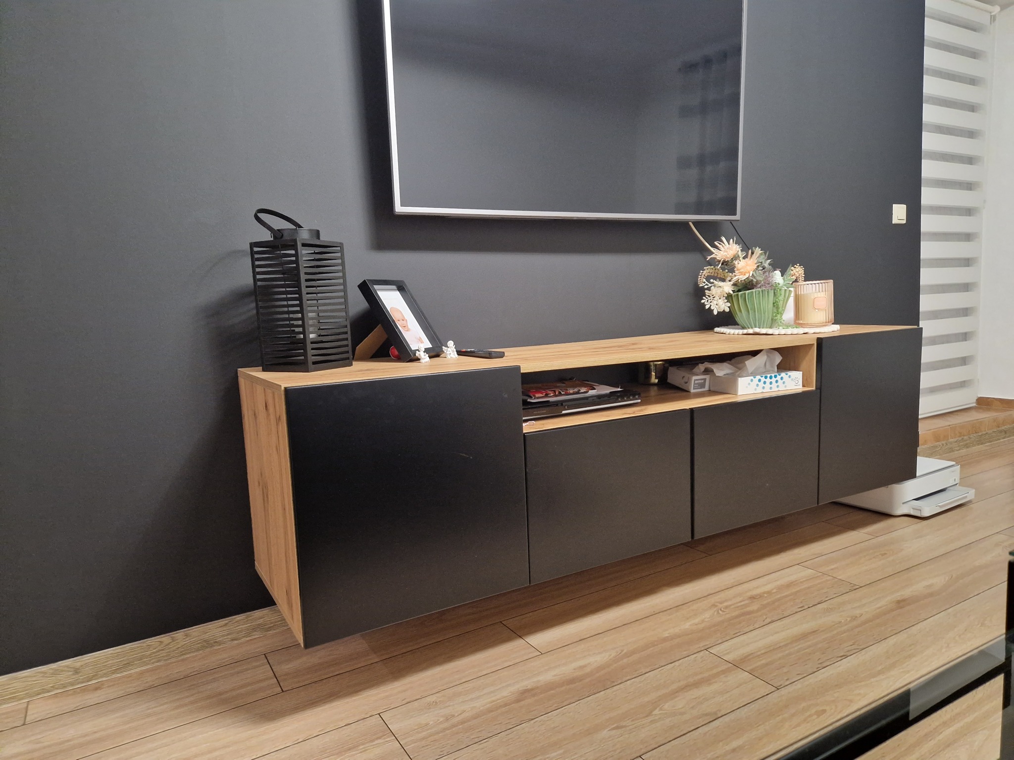Nowoczesna szafka RTV z czarnymi frontami i blatem w kolorze drewna, na której stoi dekoracja, ramka ze zdjęciem i lampion, a nad nią zawieszony telewizor na tle grafitowej ściany z roletami.