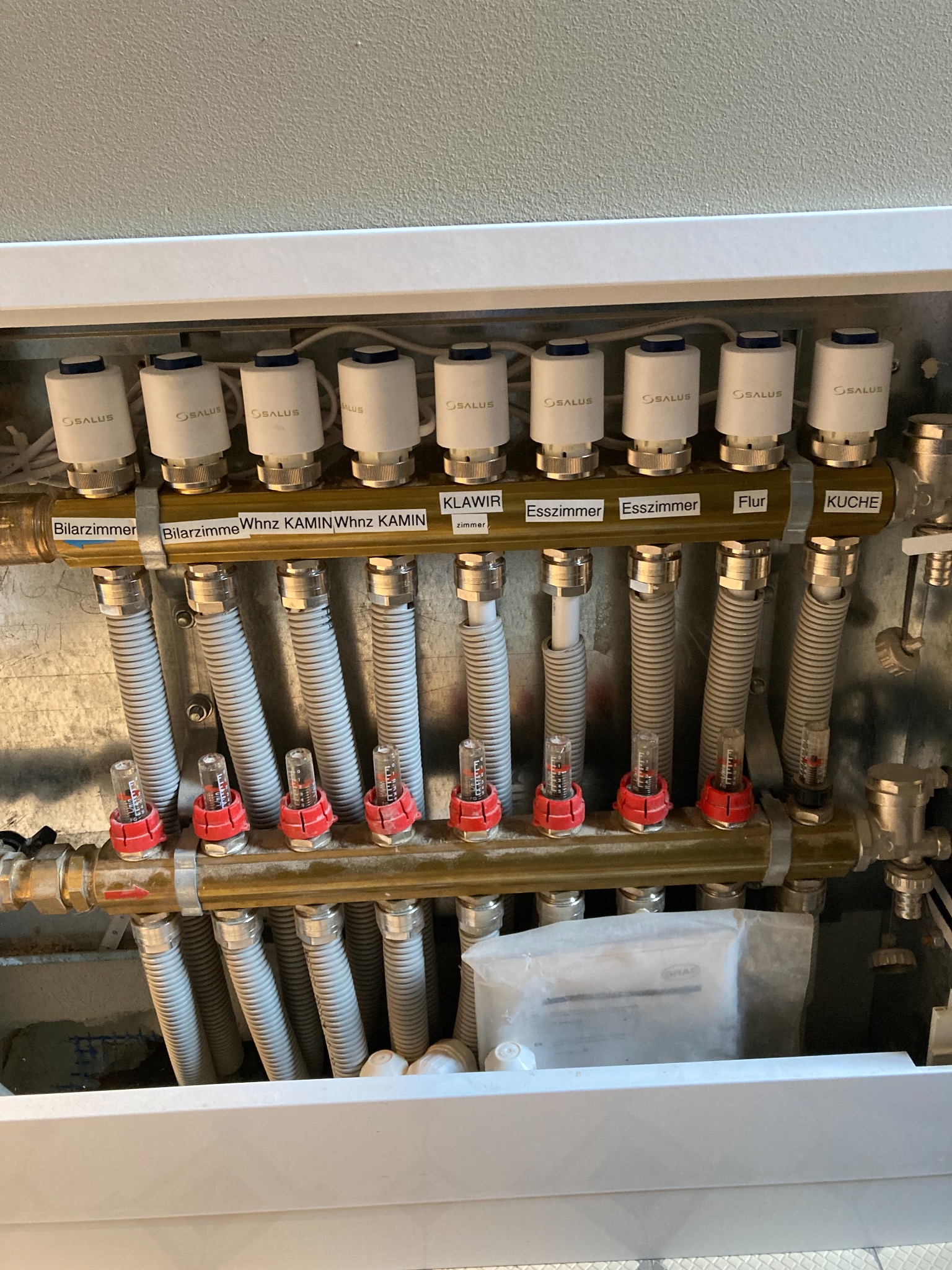 Rozdzielacz ogrzewania podłogowego z termostatami Salus, oznaczony etykietami z nazwami pomieszczeń: Bilarzimmer, Whnz KAMIN, KLAWIR, Esszimmer, Flur, KUCHE.