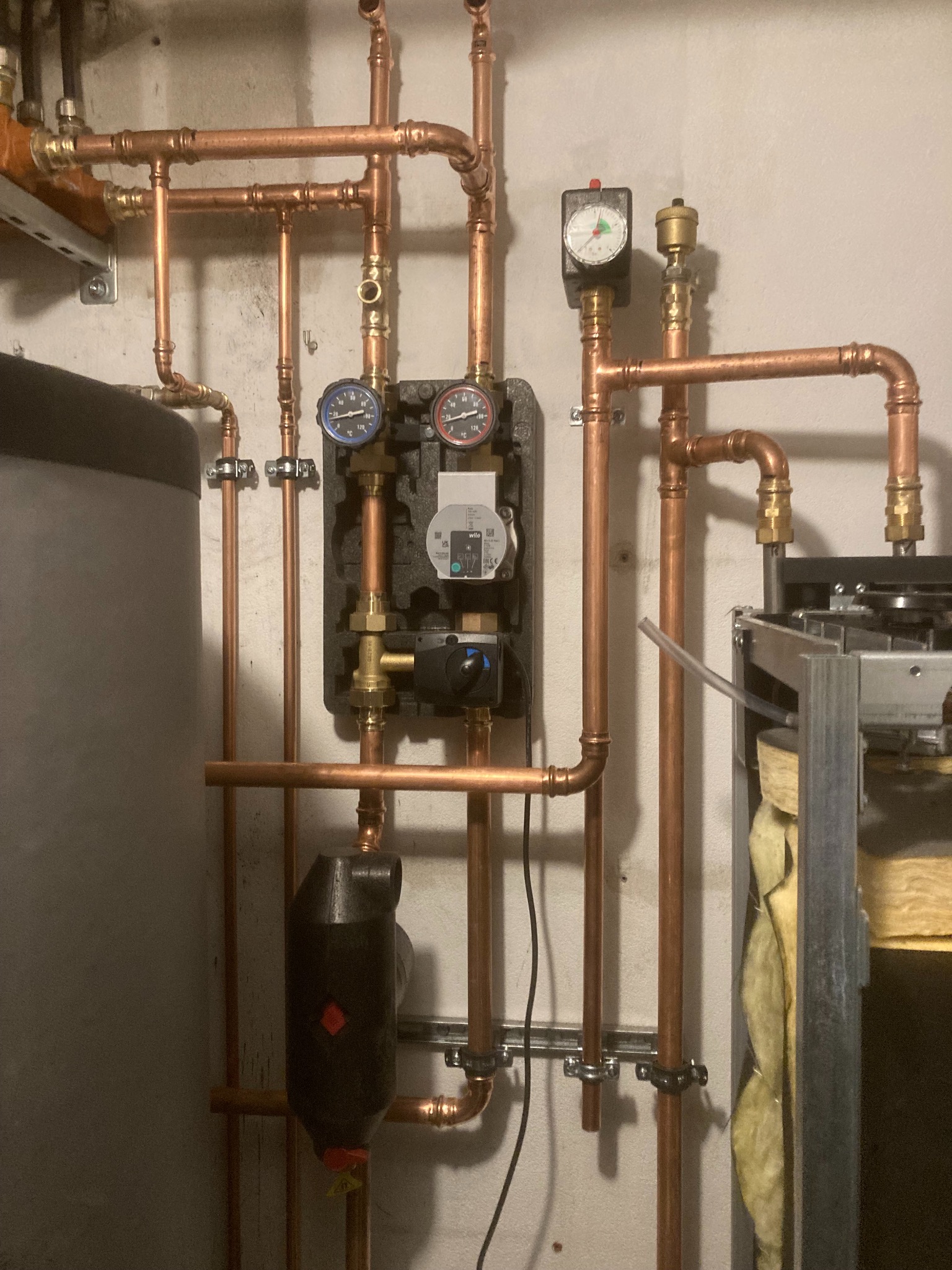 Nowa instalacja grzewcza z miedzianymi rurami, manometrami i pompą obiegową w kotłowni, widoczny zasobnik ciepłej wody i fragment pieca. Czerwone oznaczenia na czarnej obudowie pompy.