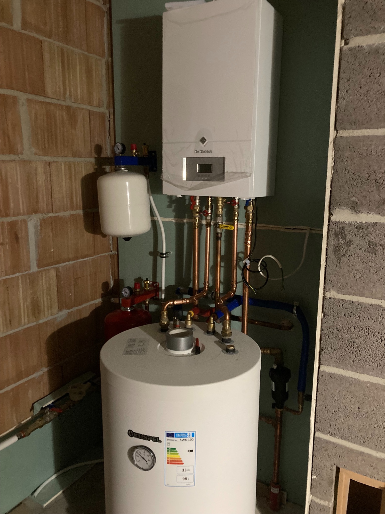 Zainstalowany, biały piec gazowy De Dietrich z podłączonym zasobnikiem Kospel SWK-100, rurami miedzianymi i elementami instalacji, widoczne manometry i etykieta energetyczna, ściana z cegły...