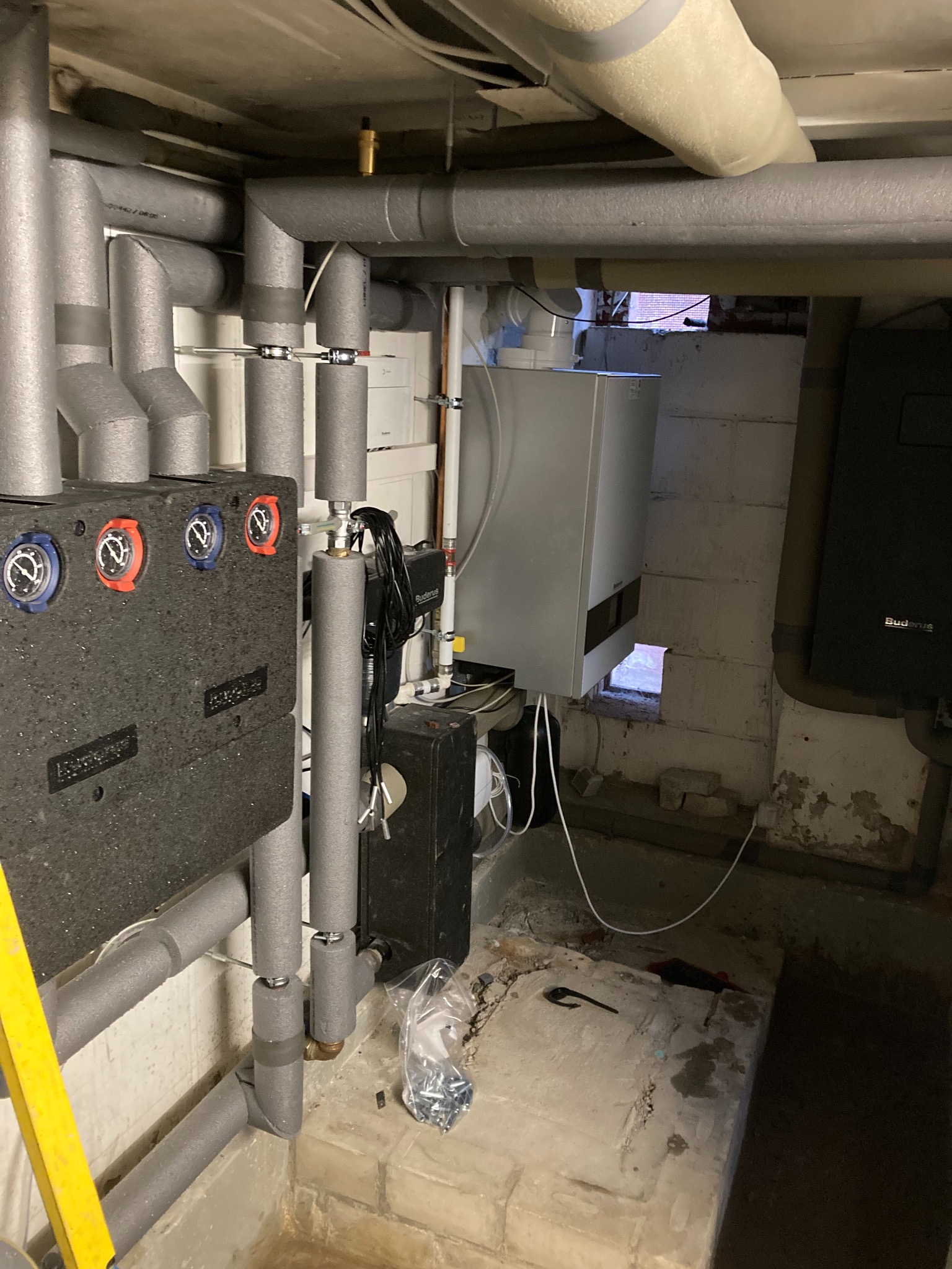 Kotłownia w piwnicy z nowym piecem gazowym, rurami grzewczymi z izolacją, osprzętem i żółtą drabiną opartą o ścianę. Widoczne ślady wilgoci na ścianach.