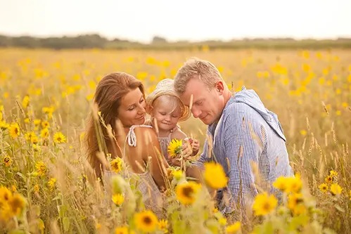 Szczęśliwa rodzina w polu słoneczników: mama trzyma córeczkę w kapeluszu, tata patrzy na kwiat w jej dłoniach, ciepłe światło zachodzącego słońca.