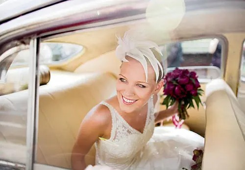 Uśmiechnięta panna młoda w koronkowej sukni i ozdobie we włosach, siedząca w beżowym wnętrzu zabytkowego samochodu z bukietem ciemnych róż.