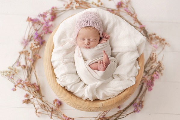 Śpiący noworodek zawinięty w biały materiał, w drewnianej misie otoczonej wiankiem z suszonych kwiatów, w różowej czapeczce.