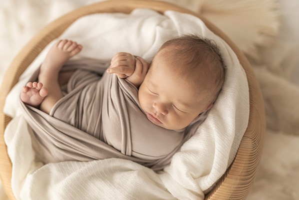 Noworodek owinięty w beżowy otulacz śpi w drewnianym koszyku na białym tle, widoczne małe stópki i rączka.