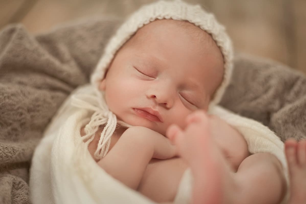 Śpiący noworodek w kremowym, dzierganym czapeczce i otulony miękkim, białym kocykiem, leżący na puszystym, beżowym tle.