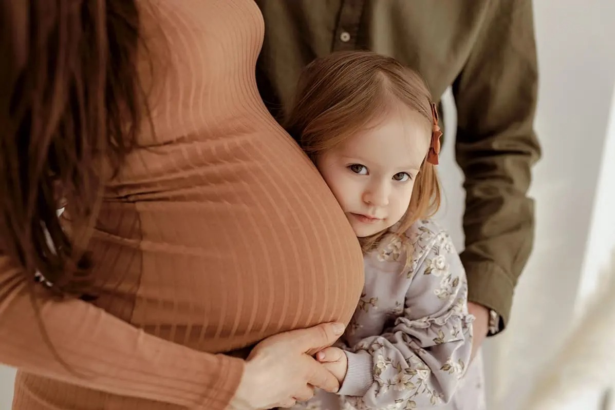 Córka przytula się do brzucha ciężarnej mamy ubranej w prążkowaną sukienkę w odcieniu terakoty; w tle widoczny mężczyzna w oliwkowej koszuli.