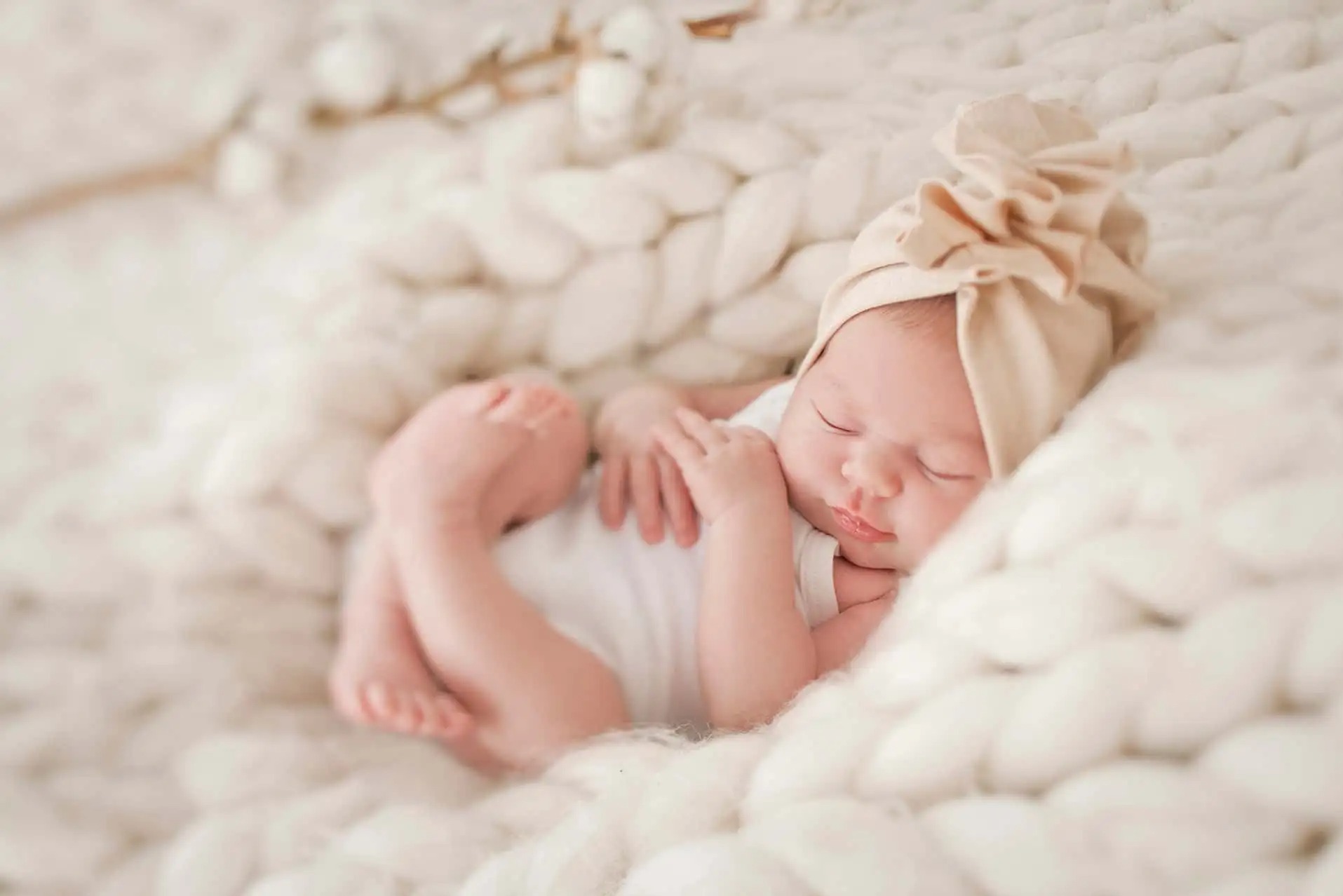 Śpiący noworodek w beżowej czapeczce z kokardą, ułożony na grubym, białym kocu z wełny czesankowej, w tle gałązka bawełny.