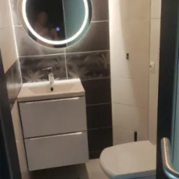 Nowoczesna łazienka z podświetlanym okrągłym lustrem, białą szafką z umywalką i toaletą; ściany wyłożone białymi i grafitowymi płytkami z motywem liści.