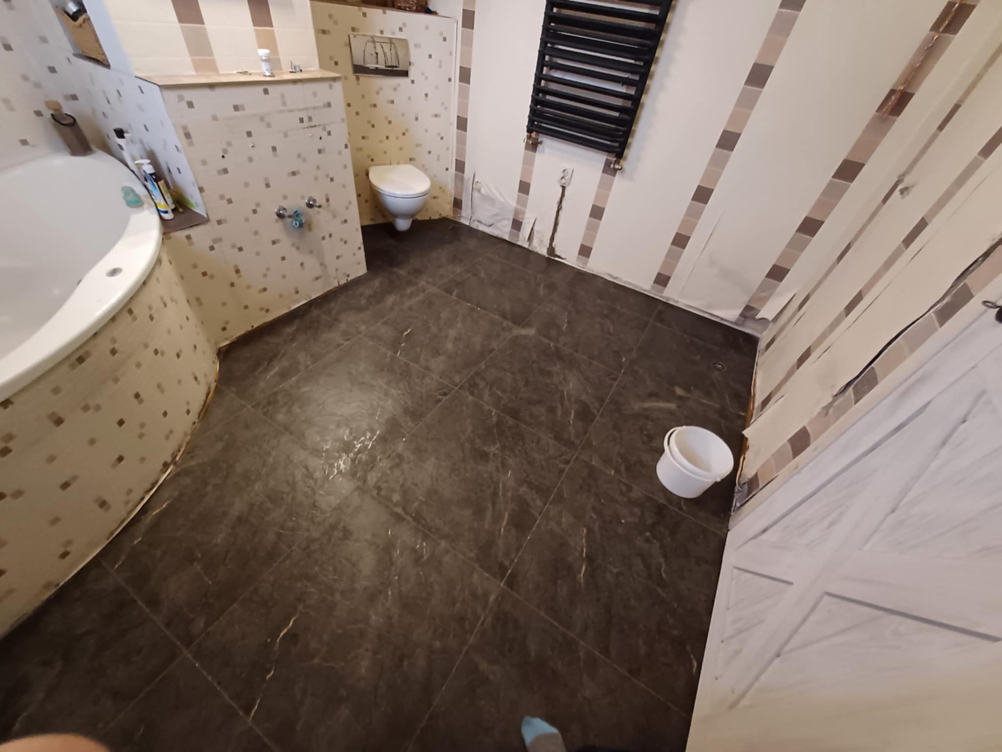 Łazienka w trakcie prac: widoczne przygotowane ściany z fragmentami odklejonej taśmy malarskiej, ciemna podłoga z płytek imitujących kamień, białe sanitariaty oraz grzejnik drabinkowy. W rogu stoi...