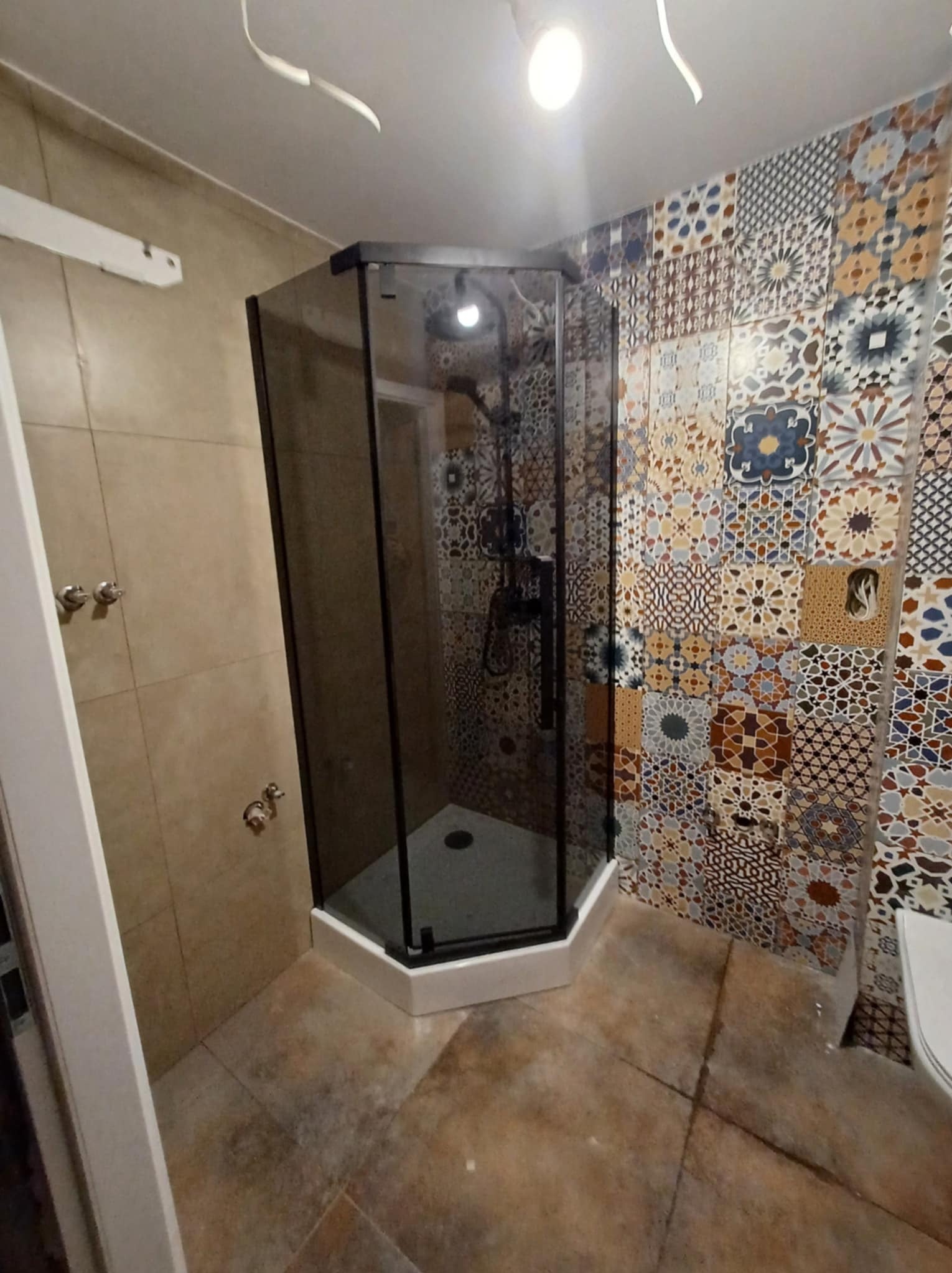 Kabina prysznicowa z czarnymi profilami w rogu łazienki, ściana wyłożona patchworkiem płytek w stylu marokańskim, beżowe płytki na pozostałych ścianach i podłodze.