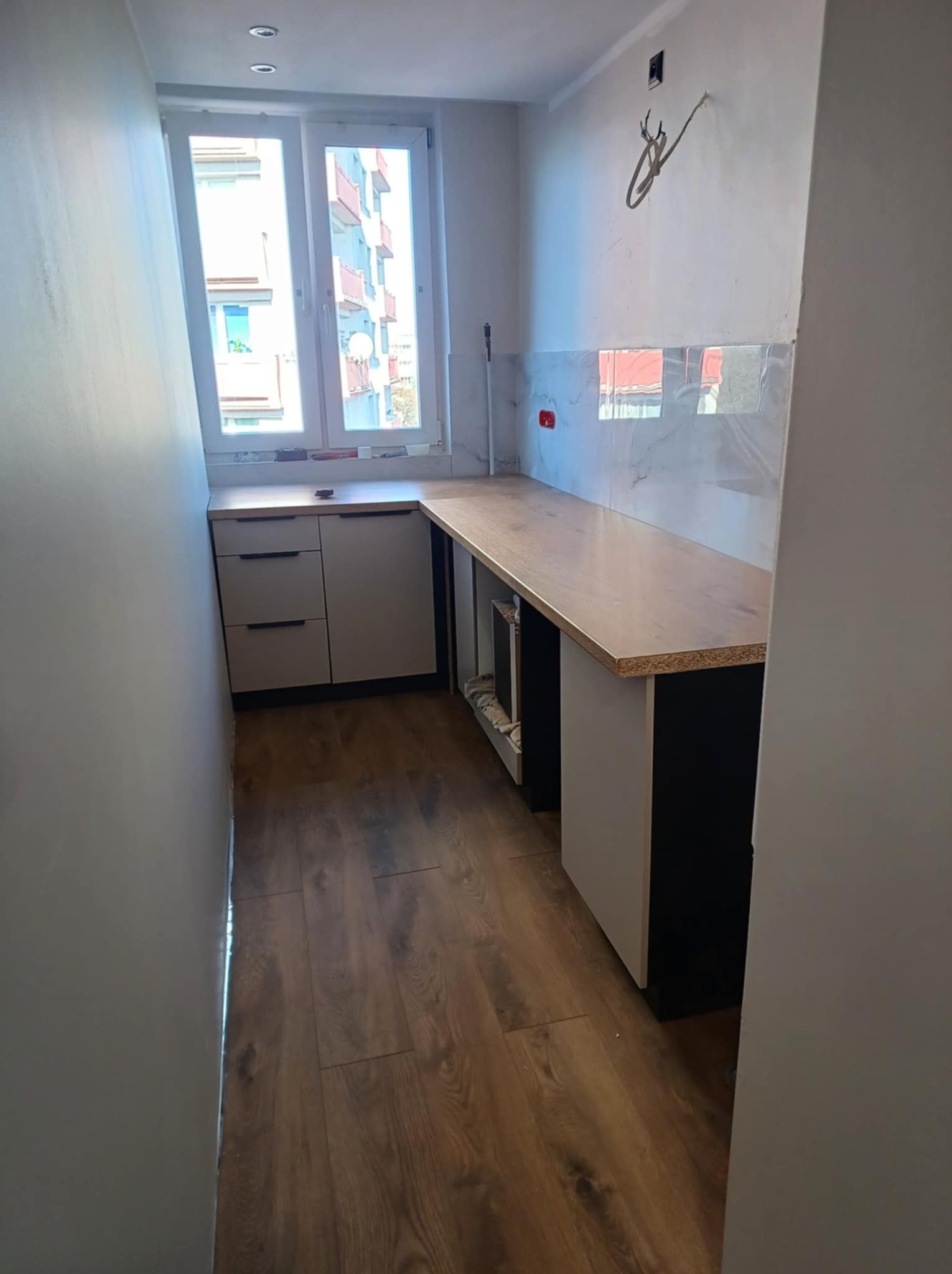 Wnętrze kuchni w trakcie wykańczania: widoczne szafki kuchenne z blatem, częściowo położona glazura imitująca marmur na ścianie, drewnopodobna podłoga, okno z widokiem na budynek mieszkalny...