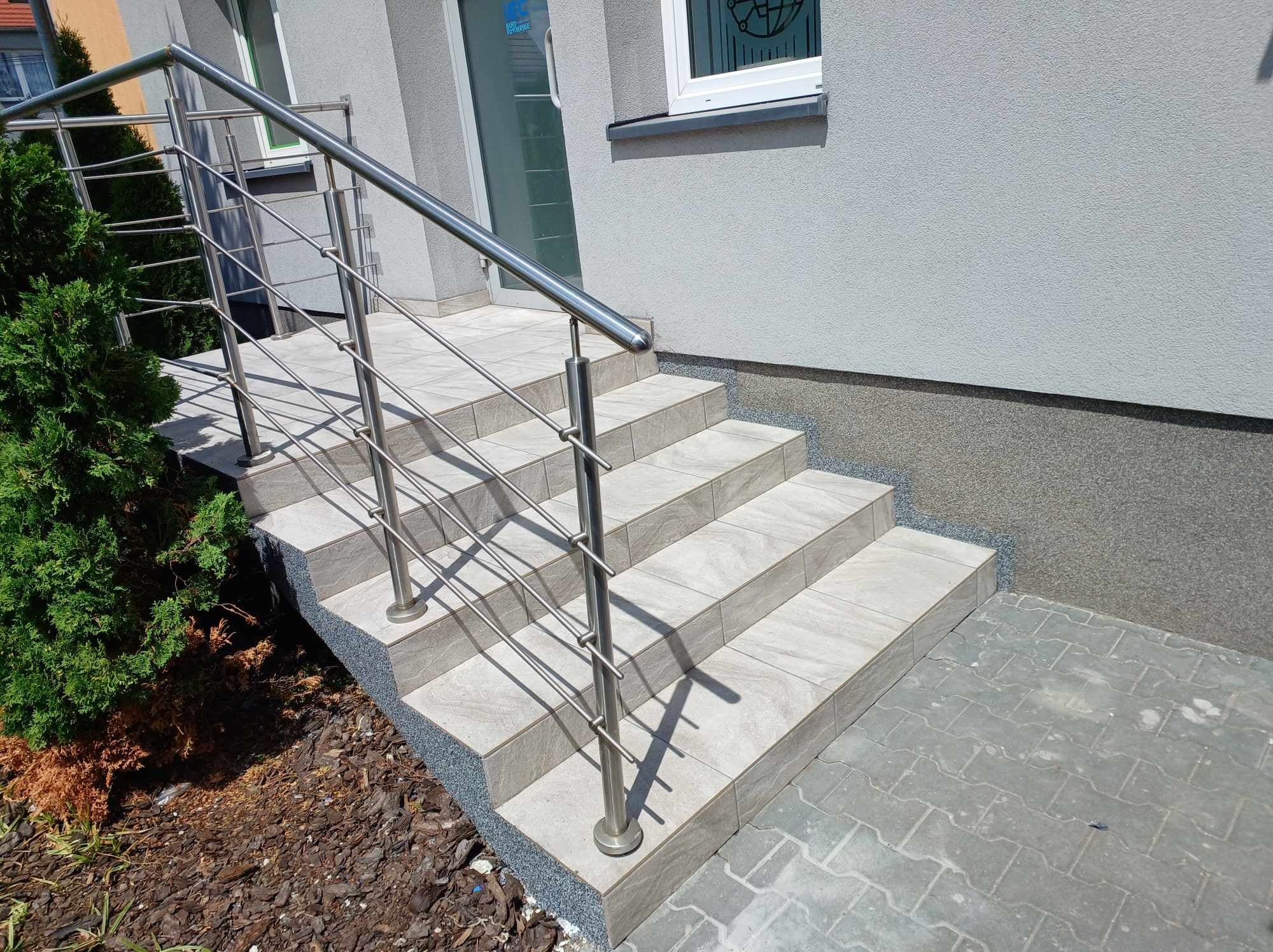 Zewnętrzne schody wejściowe wyłożone jasnymi płytkami ceramicznymi z metalową balustradą, widok z dołu, z boku rośnie krzew iglasty.