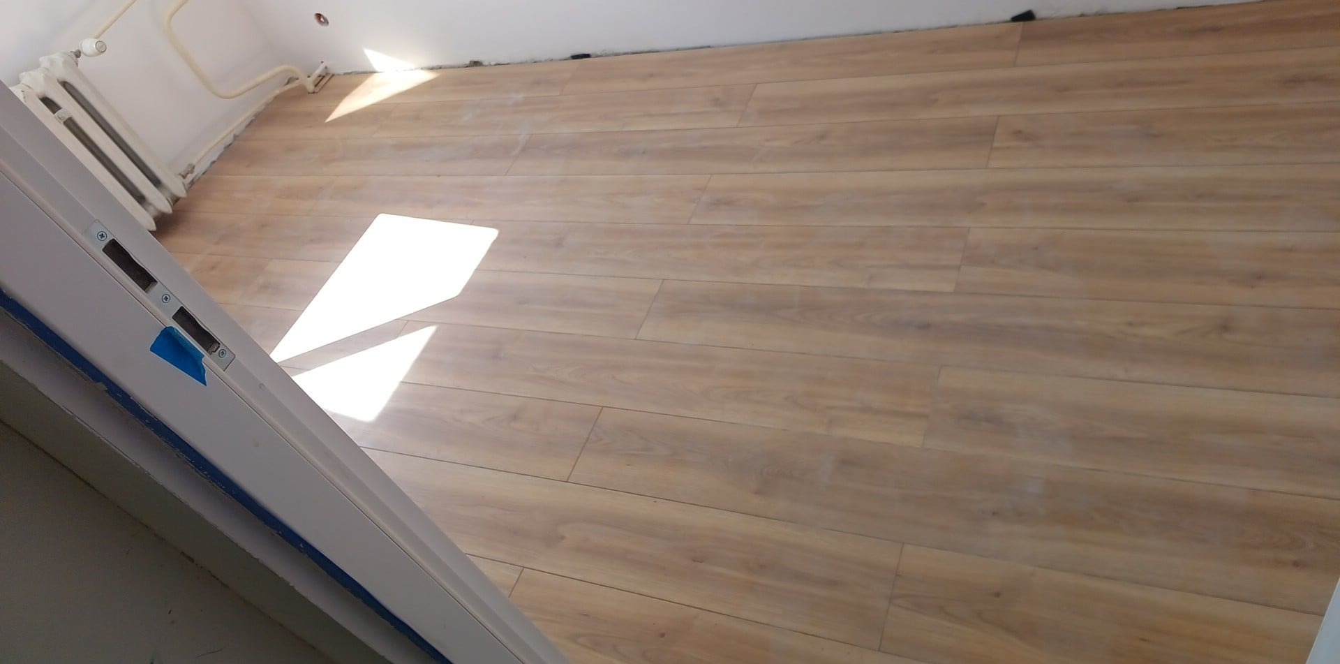 Ułożona podłoga z paneli drewnopodobnych w jasnym odcieniu, widoczny fragment drzwi z niebieską taśmą zabezpieczającą oraz grzejnik żeliwny przy ścianie.