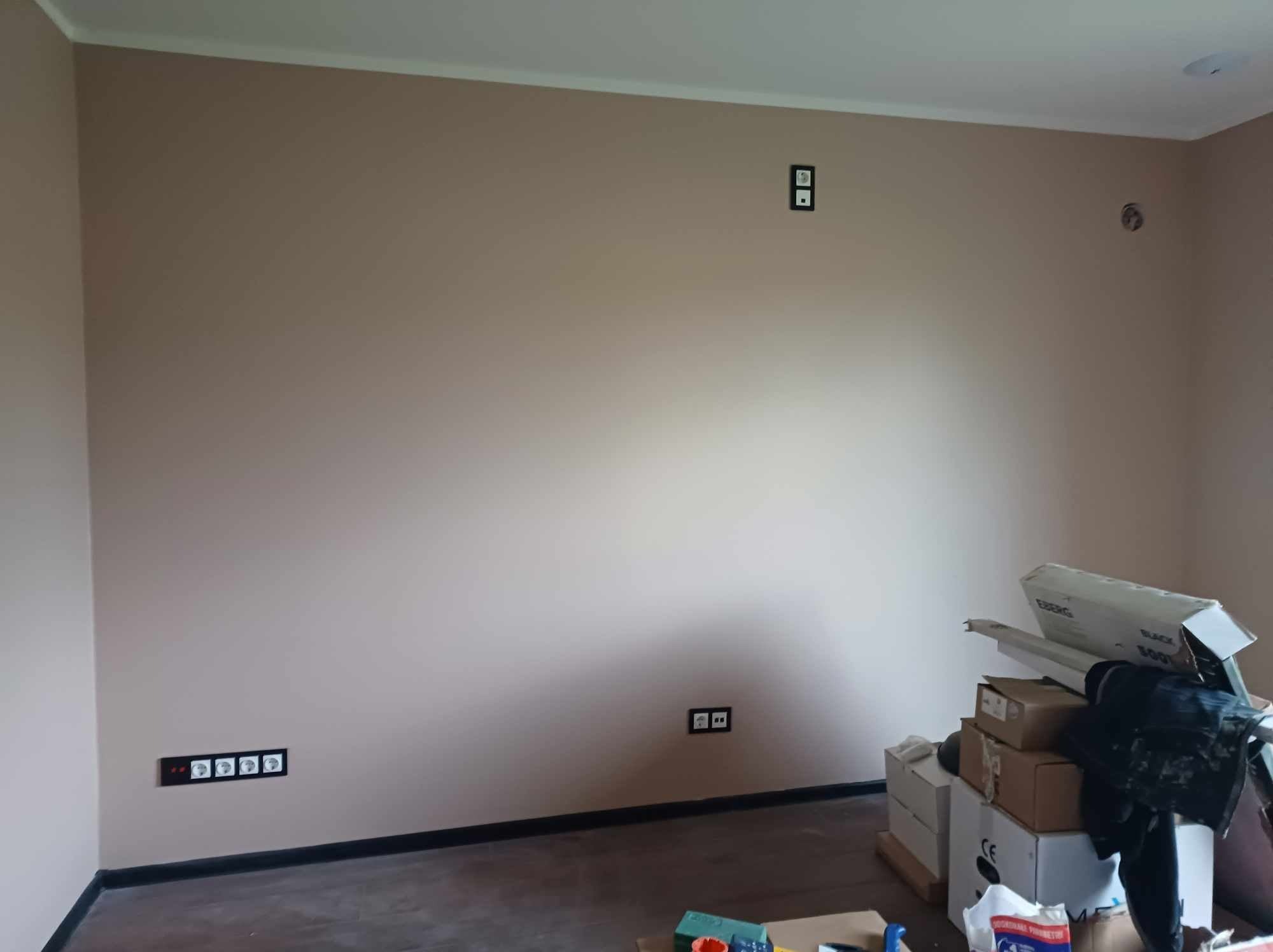 Ściana w odcieniach beżu z zamontowanymi gniazdkami elektrycznymi w czarnych ramkach, widoczne również kartony i materiały budowlane w rogu pomieszczenia.