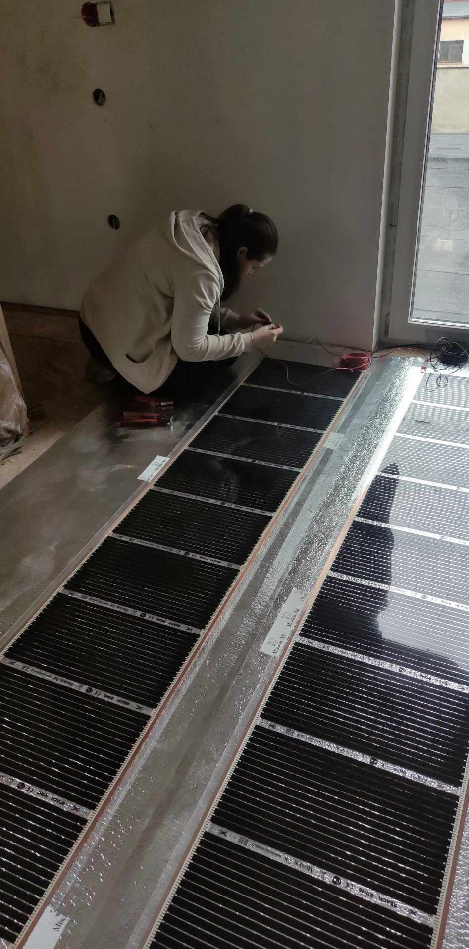 Kobieta w jasnej bluzie łączy przewody do mat grzewczych rozłożonych na podłodze w surowym stanie, w pobliżu okna i gniazdek elektrycznych.