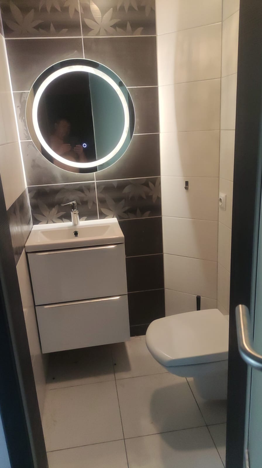 Nowoczesna łazienka z podświetlanym okrągłym lustrem, białą szafką z umywalką i toaletą; ściany wyłożone białymi i grafitowymi płytkami z motywem liści.