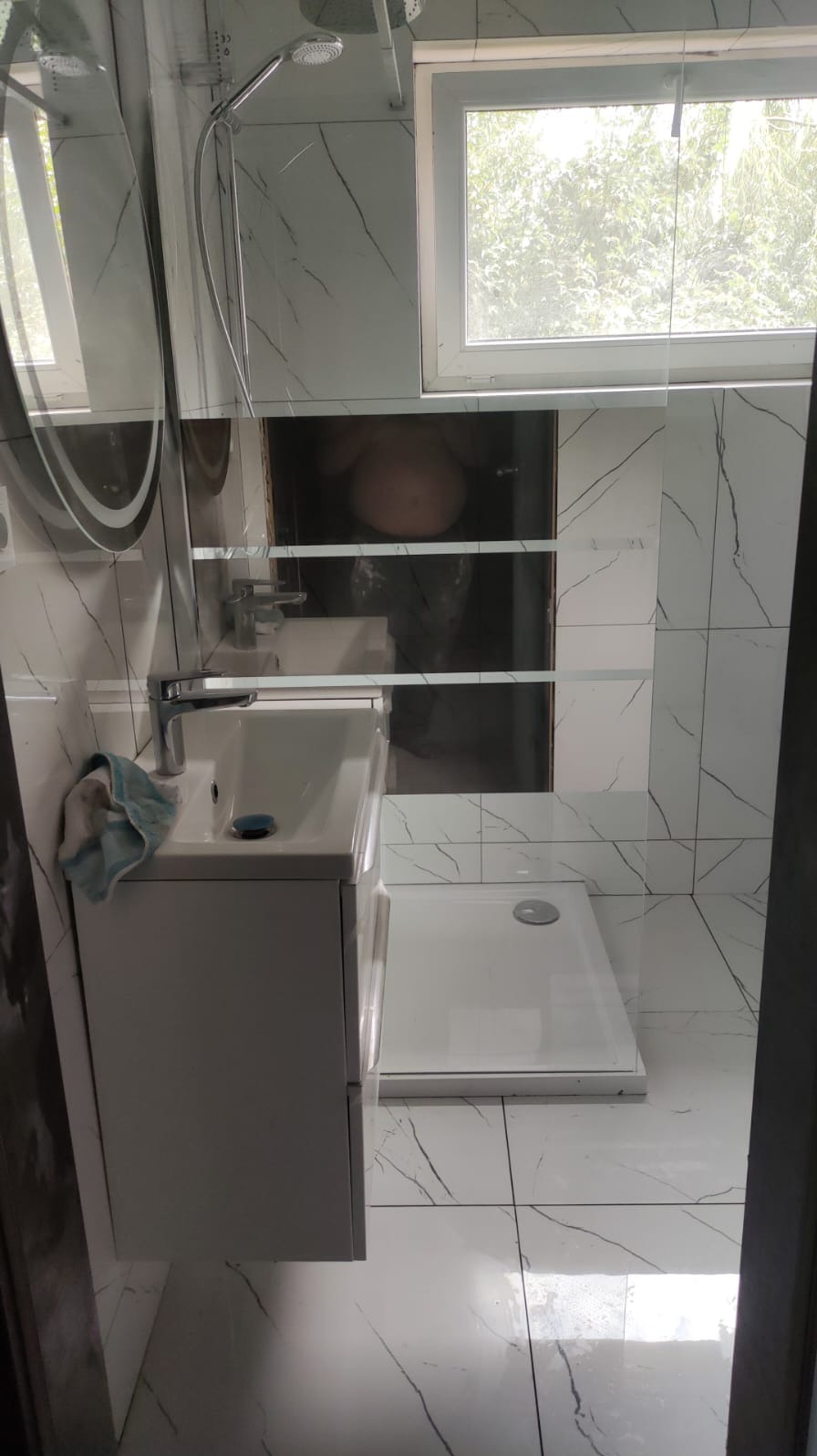 Nowoczesna łazienka z białą umywalką nablatową, kabiną prysznicową i marmurowymi płytkami; widoczne niedokończone prace w postaci niezamaskowanego otworu w ścianie.