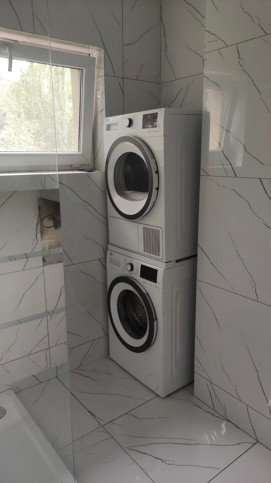 Ustawiona pionowo pralka i suszarka w wykończonej łazience z białymi płytkami imitującymi marmur, widok na okno z boku.
