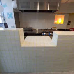 Dawid Flig - Biała, kafelkowana lada z żółtymi fugami w lokalu gastronomicznym. W tle widoczny sprzęt kuchenny i okap. Minimalistyczny design.