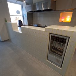 Dawid Flig - Biała, kafelkowana lada z żółtymi fugami w lokalu gastronomicznym. Wbudowana lodówka wypełniona napojami. W tle okap i kafelkowana ściana.