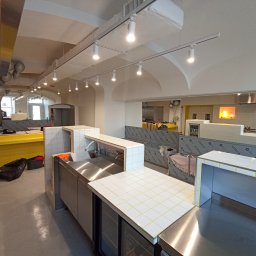 Dawid Flig - Wnętrze lokalu gastronomicznego w trakcie wykańczania. Widoczne metalowe blaty i elementy wykończone białymi płytkami z żółtą fugą, surowy strop z oświetleniem.