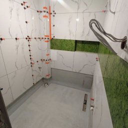 Dawid Flig - Wnętrze łazienki w trakcie kładzenia białych płytek z szarym wzorem, z użyciem systemu poziomowania płytek. Widoczny fragment ściany z zielonym mchem.