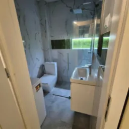 Nowoczesna łazienka z marmurowymi płytkami, szklaną kabiną prysznicową, białą umywalką z szafką i toaletą. Widok zza uchylonych drzwi.