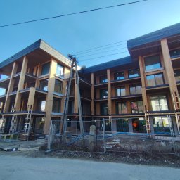 Dawid Flig - Wielopiętrowy budynek w trakcie budowy z drewnianą konstrukcją szkieletową i zamontowanymi oknami, otoczony tymczasowym ogrodzeniem i rusztowaniami.