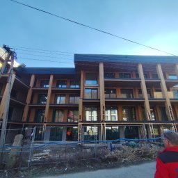 Dawid Flig - Wielopiętrowy budynek z drewnianą elewacją i dużymi oknami, w trakcie budowy. Widoczne rusztowania i ogrodzenie budowy. Błękitne niebo w tle.