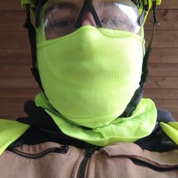 Dawid Flig - Portret mężczyzny w kasku ochronnym, goglach i masce ochronnej na tle drewnianej ściany. Widoczne elementy odzieży roboczej i logo na kasku.