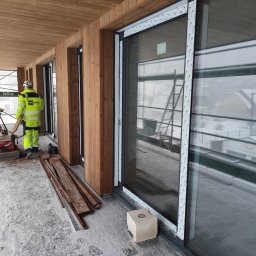 Dawid Flig - Montaż dużych okien przesuwnych z drewnianą elewacją. Widoczny pracownik w odblaskowej kurtce i piła do cięcia drewna na zewnątrz budynku w zimowych warunkach.