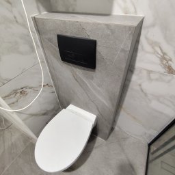 Dawid Flig - Kompaktowa toaleta podwieszana z czarnym przyciskiem spłukującym na tle szarej, marmurkowej ściany w nowoczesnej łazience. Widoczne płytki podłogowe w odcieniach szarości.
