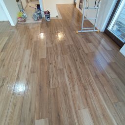 Dawid Flig - Połyskująca, świeżo ułożona podłoga z płytek imitujących drewno. Widoczne narzędzia glazurnicze i drabina. Jasne wnętrze w trakcie prac wykończeniowych.