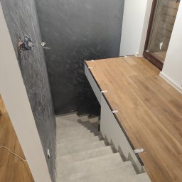 Dawid Flig - Wykończenie schodów z drewnopodobnymi płytkami na podeście i szarymi stopnicami. Dekoracyjna szara ściana. Widoczne elementy instalacji elektrycznej.
