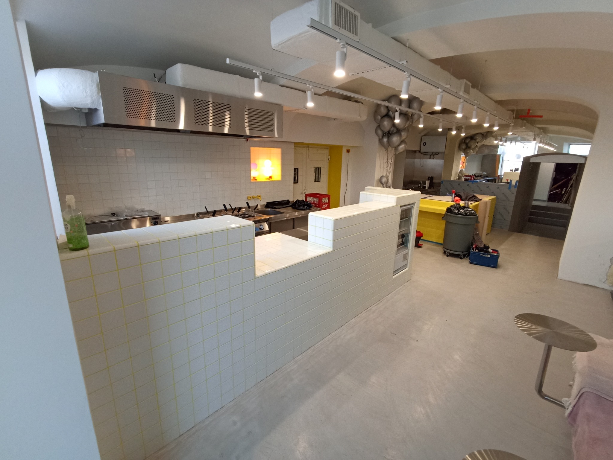 Biała, kafelkowana lada w lokalu gastronomicznym z widoczną kuchnią i elementami wyposażenia. Widok na fragment sali z balonami i stolikami. Jasne oświetlenie.