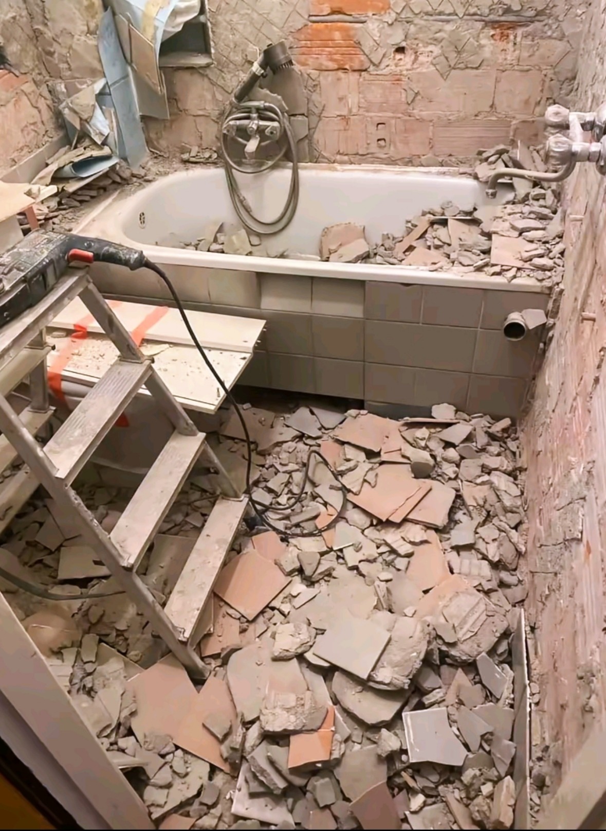 Demontaż płytek w łazience. Widoczna wanna, drabina, gruz i narzędzie do skuwania płytek. Etap remontu z bliska, stare płytki usunięte ze ścian i podłogi.