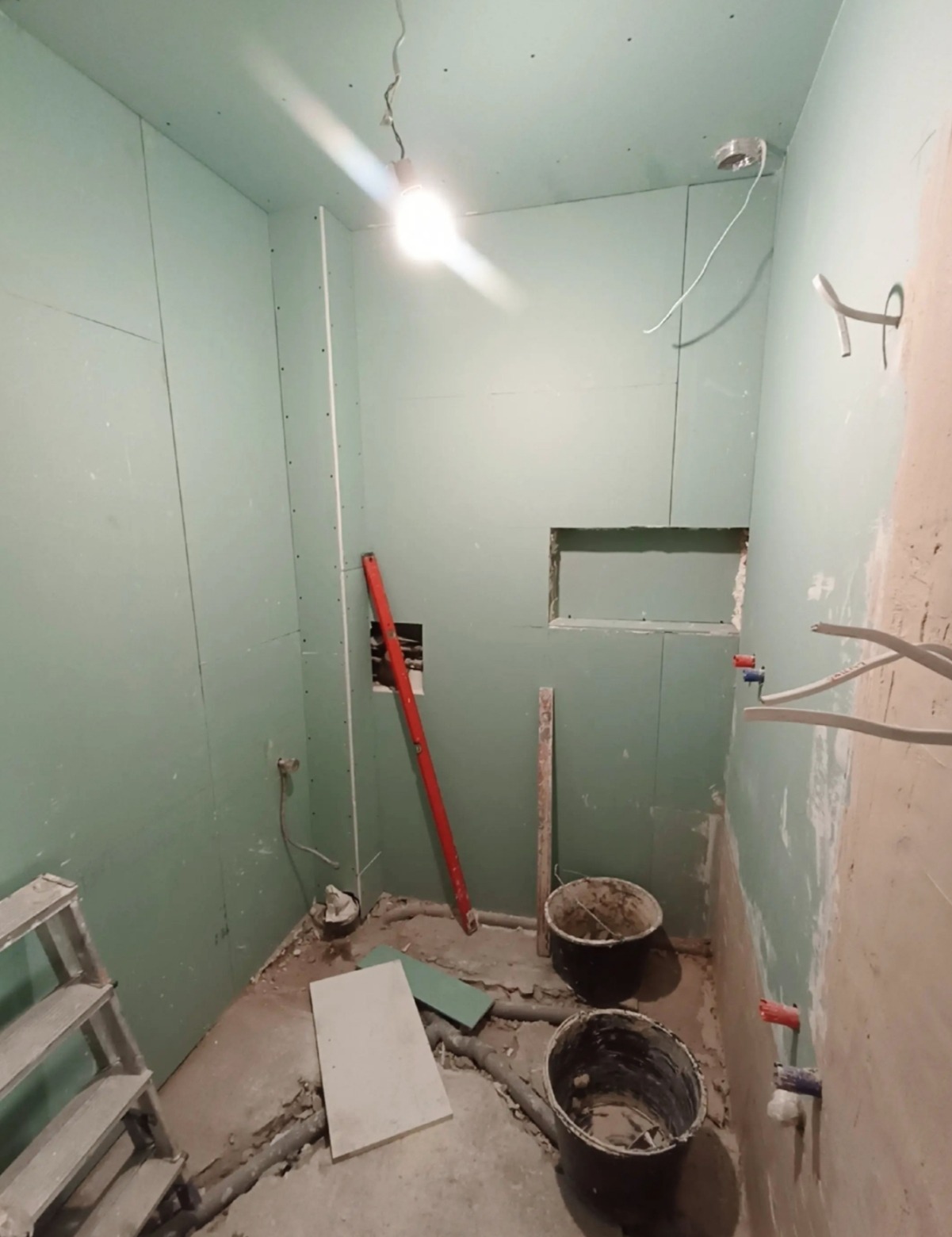 Łazienka w trakcie remontu: szpachlowane ściany, płyty gipsowo-kartonowe, poziomica oparta o ścianę, dwa wiadra i drabina w pomieszczeniu. Widoczne instalacje.