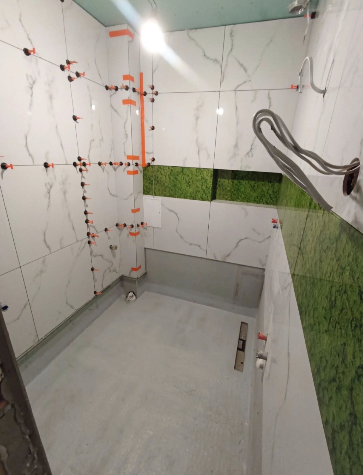 Wnętrze łazienki w trakcie kładzenia białych płytek z szarym wzorem, z użyciem systemu poziomowania płytek. Widoczny fragment ściany z zielonym mchem.