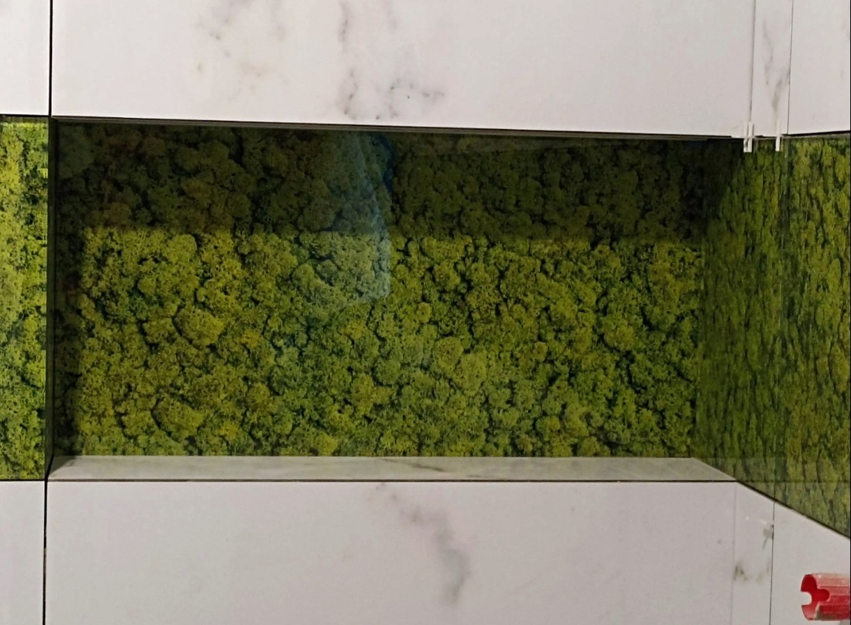 Ściana z białych płytek marmurowych z wnęką wypełnioną zielonym mchem. Dekoracja wnętrza z użyciem naturalnych elementów.