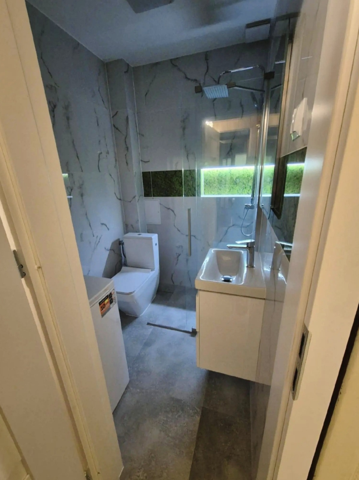 Nowoczesna łazienka z marmurowymi płytkami, szklaną kabiną prysznicową, białą umywalką z szafką i toaletą. Widok zza uchylonych drzwi.