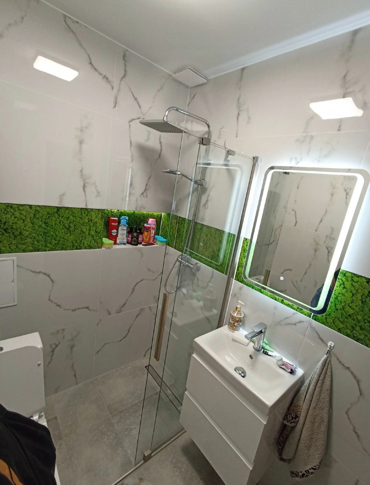 Wykończona łazienka z kabiną prysznicową typu walk-in, umywalką na szafce i lustrem z podświetleniem LED. Ściany wyłożone płytkami imitującymi marmur, akcent zielonego mchu.