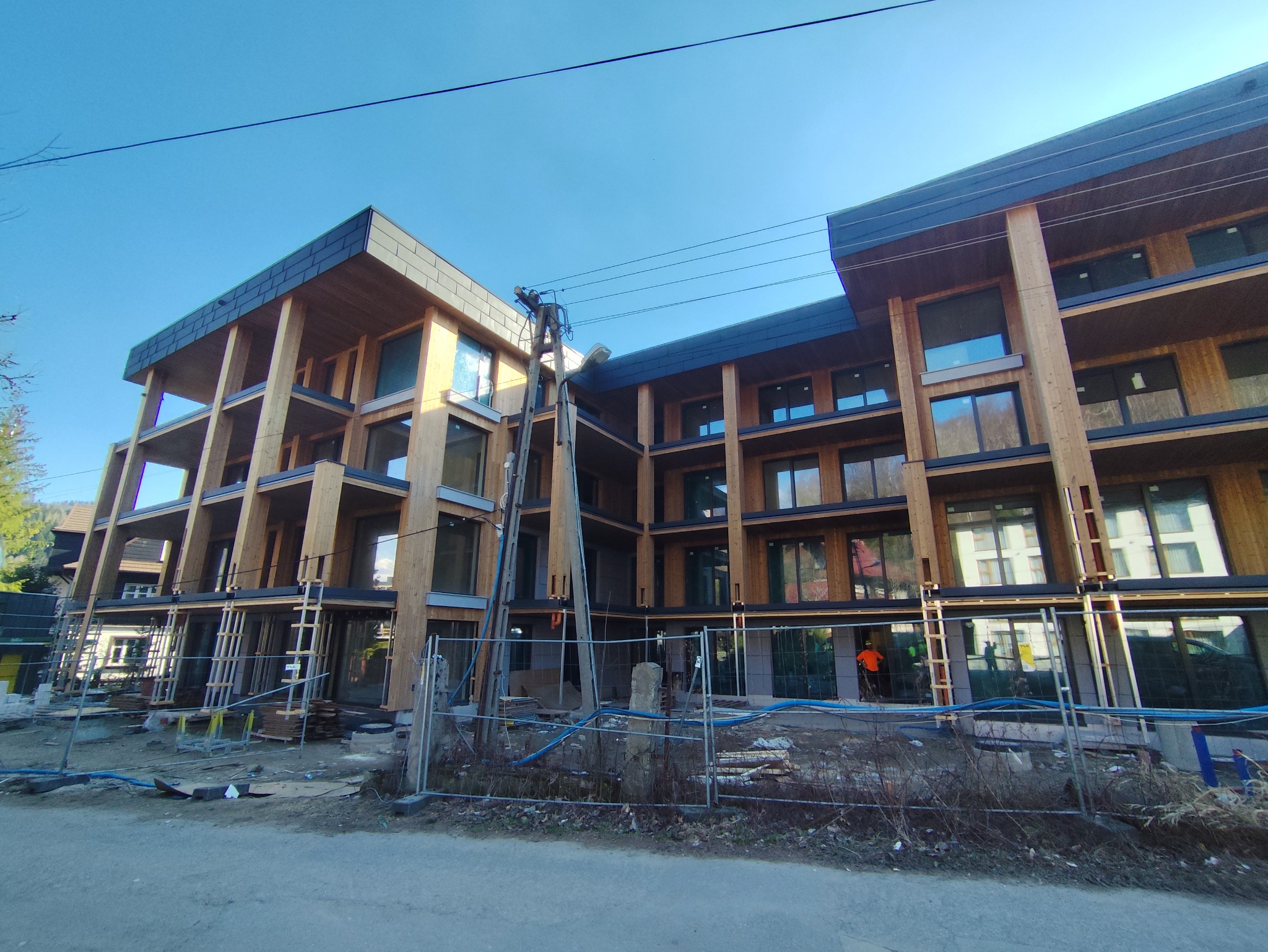 Wielopiętrowy budynek w trakcie budowy z drewnianą konstrukcją szkieletową i zamontowanymi oknami, otoczony tymczasowym ogrodzeniem i rusztowaniami.