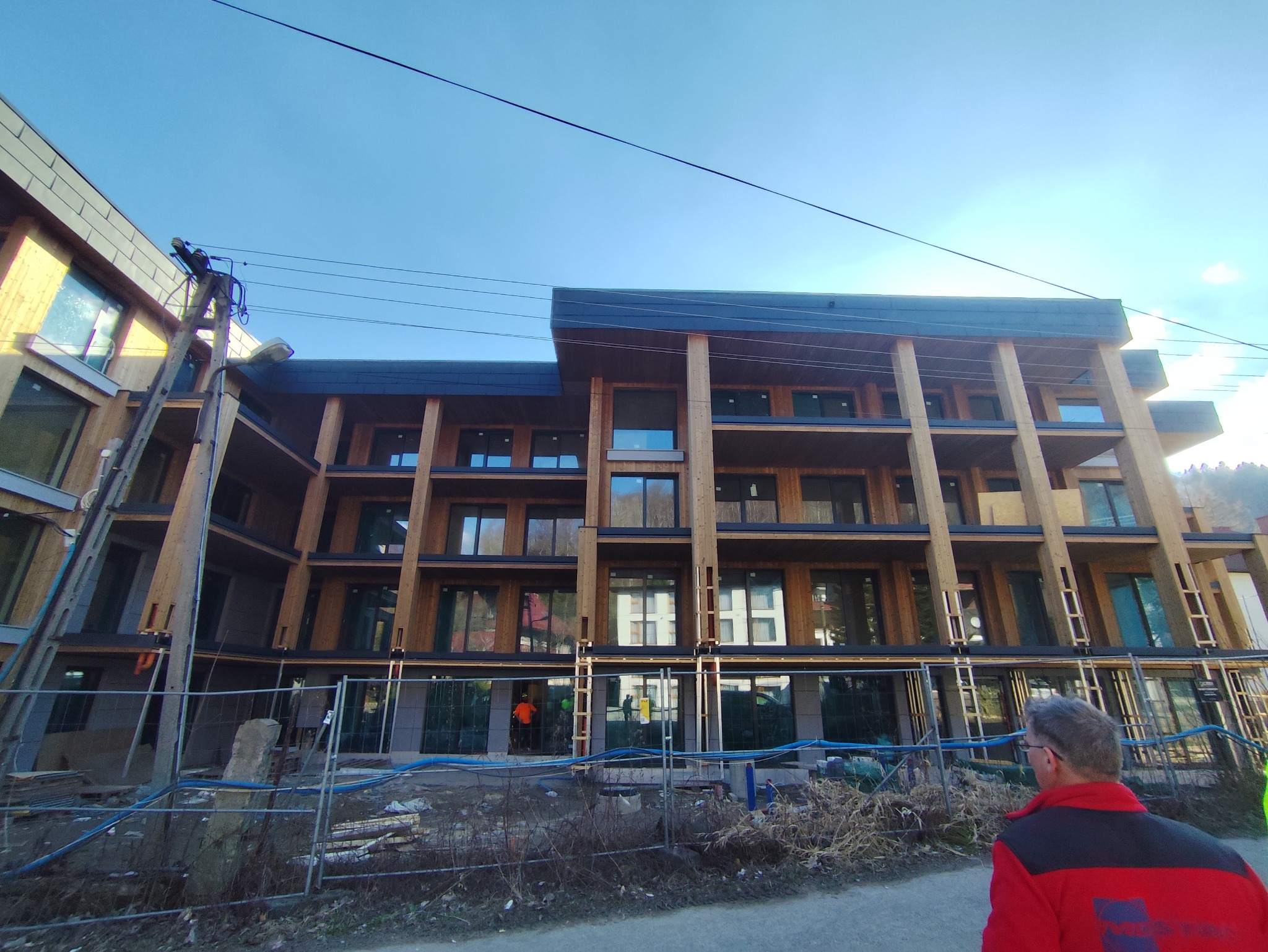 Wielopiętrowy budynek z drewnianą elewacją i dużymi oknami, w trakcie budowy. Widoczne rusztowania i ogrodzenie budowy. Błękitne niebo w tle.