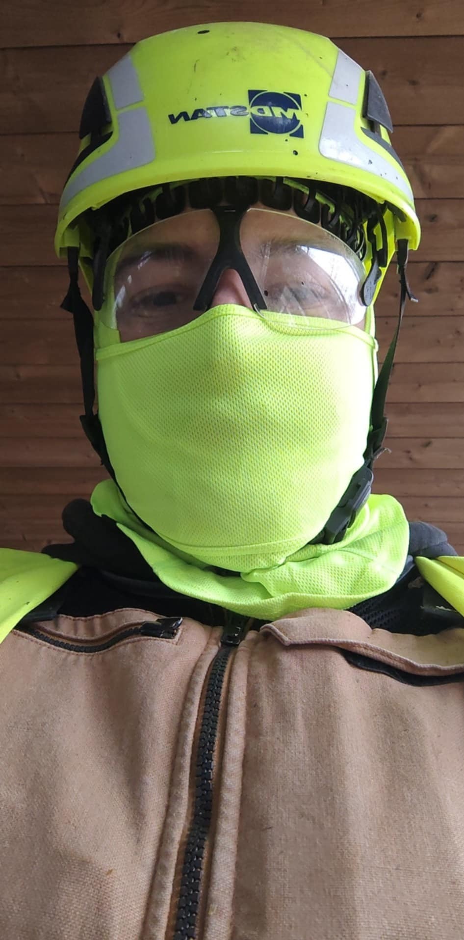 Portret mężczyzny w kasku ochronnym, goglach i masce ochronnej na tle drewnianej ściany. Widoczne elementy odzieży roboczej i logo na kasku.