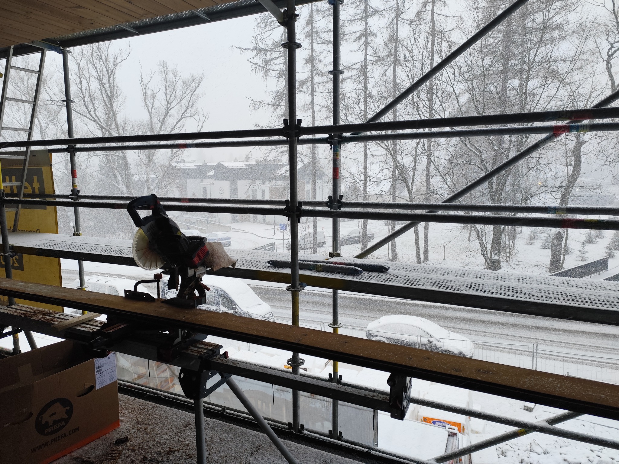 Zimowy kadr z rusztowania: piła do cięcia, deski i zasypane śniegiem otoczenie. W tle budynki i drzewa w śnieżnej scenerii.