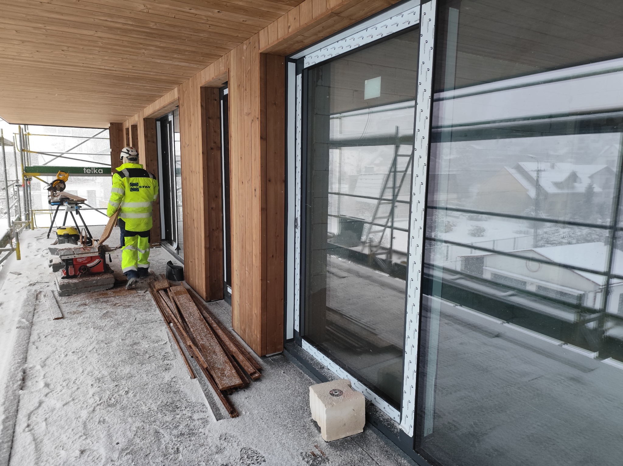 Montaż dużych okien przesuwnych z drewnianą elewacją. Widoczny pracownik w odblaskowej kurtce i piła do cięcia drewna na zewnątrz budynku w zimowych warunkach.