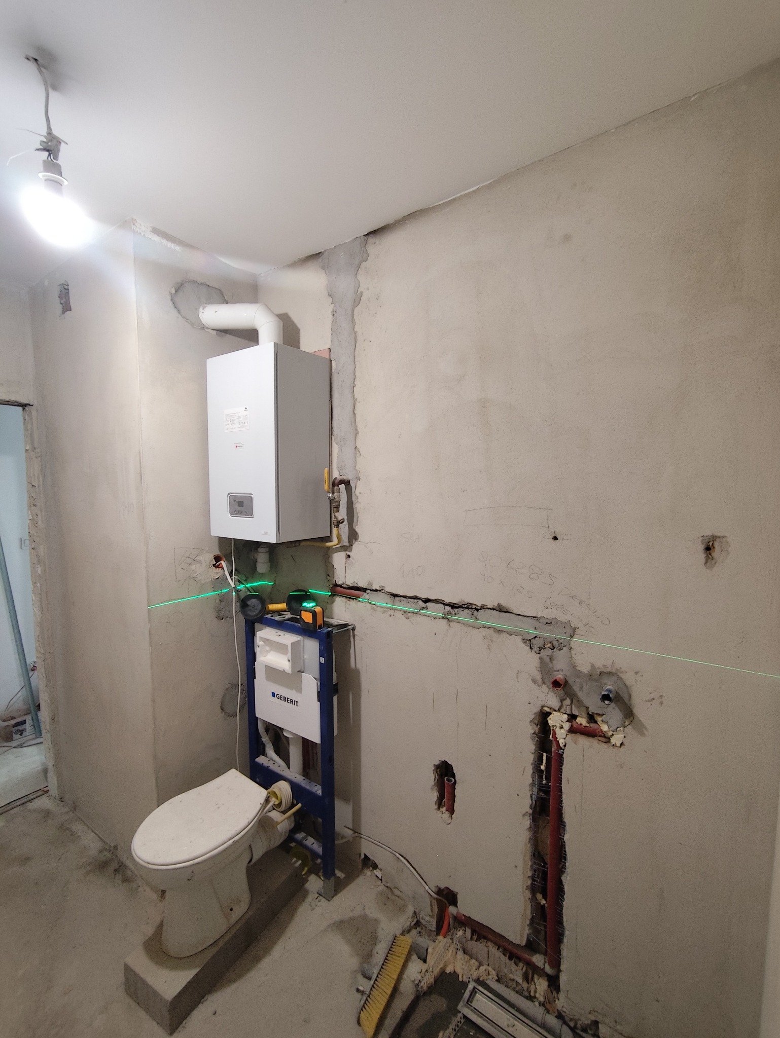 Niedokończona łazienka z zamontowanym stelażem WC Geberit, piecem gazowym i widocznymi instalacjami. Użyto poziomicy laserowej. Ściany surowe, przygotowane do dalszych prac.