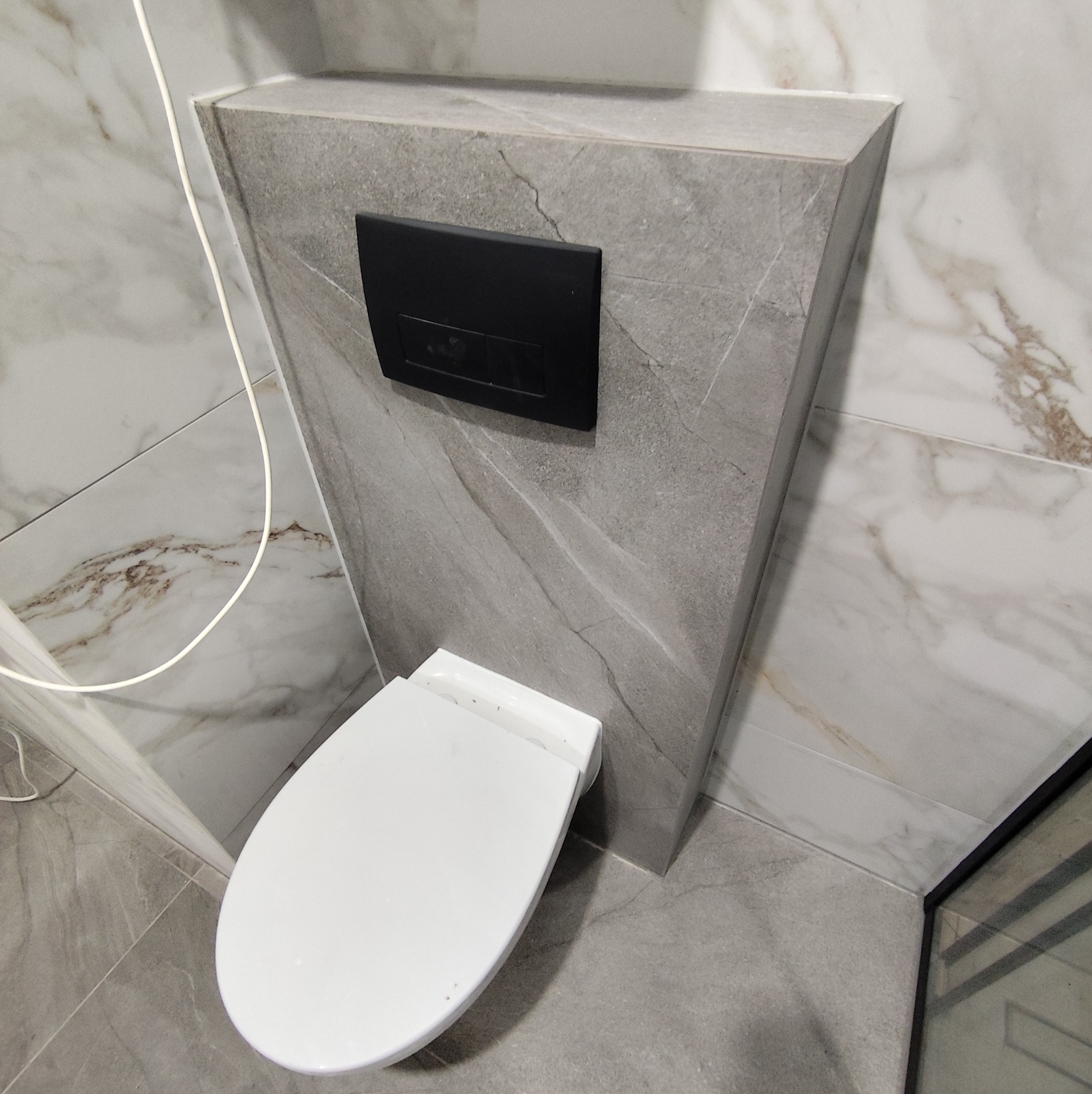 Kompaktowa toaleta podwieszana z czarnym przyciskiem spłukującym na tle szarej, marmurkowej ściany w nowoczesnej łazience. Widoczne płytki podłogowe w odcieniach szarości.